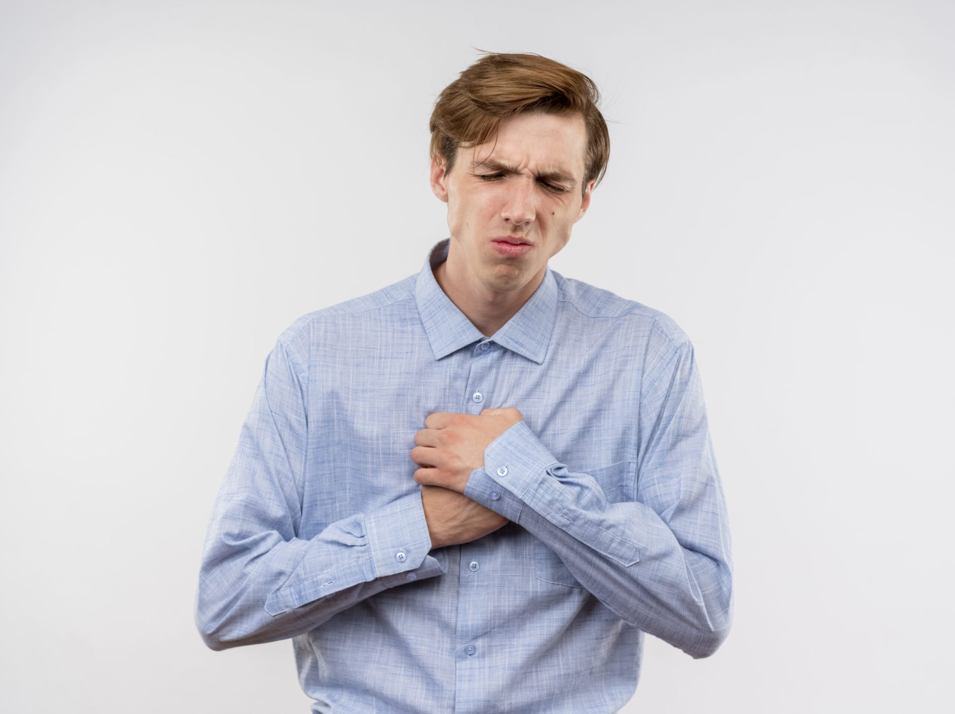 young man blue shirt holding hnads chest looking unwell feeling pain standing white wall scaled 1